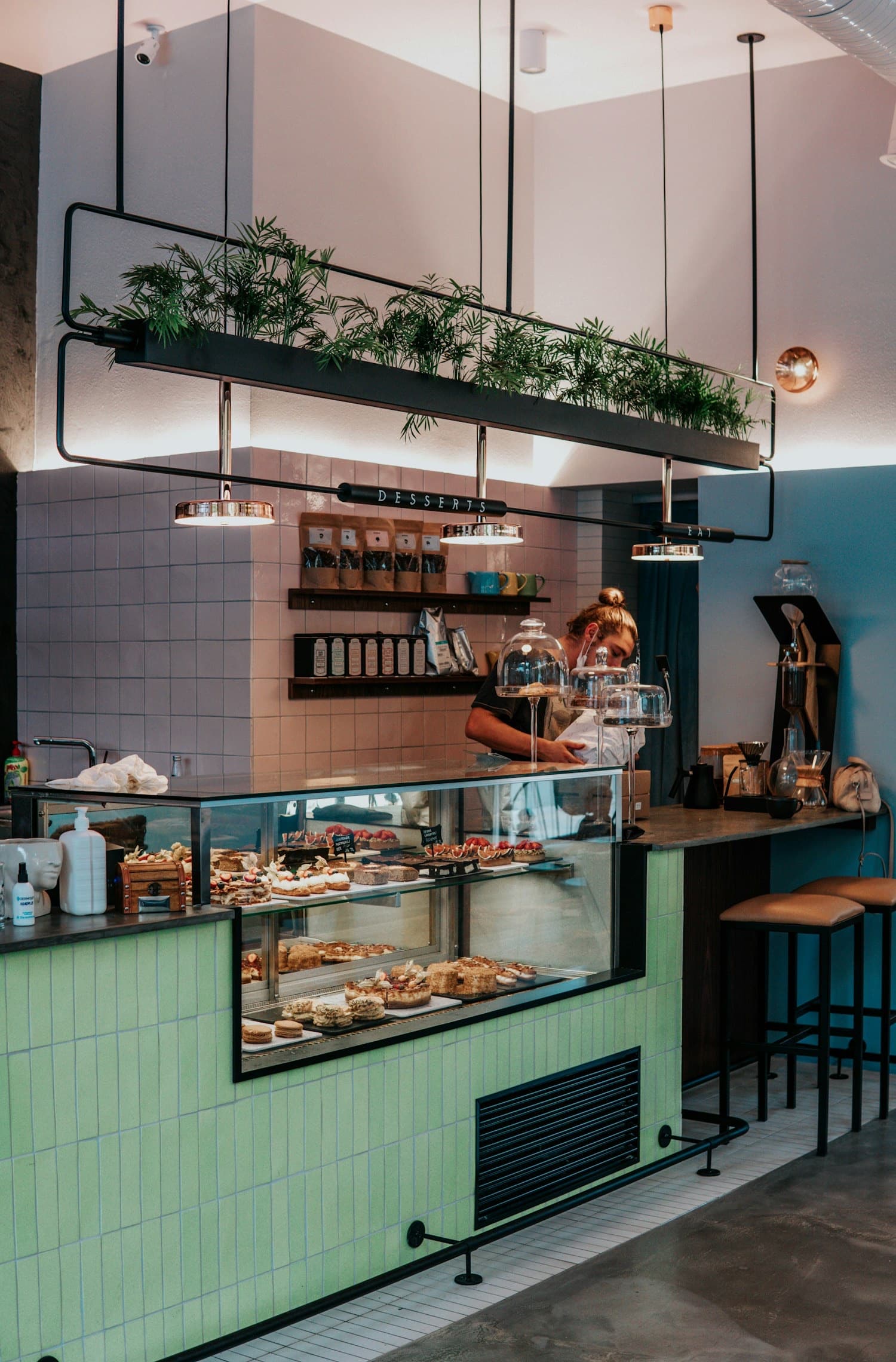 Comptoir pastel d'un café : carrelage menthe, vitrine de pâtisseries et plantes.